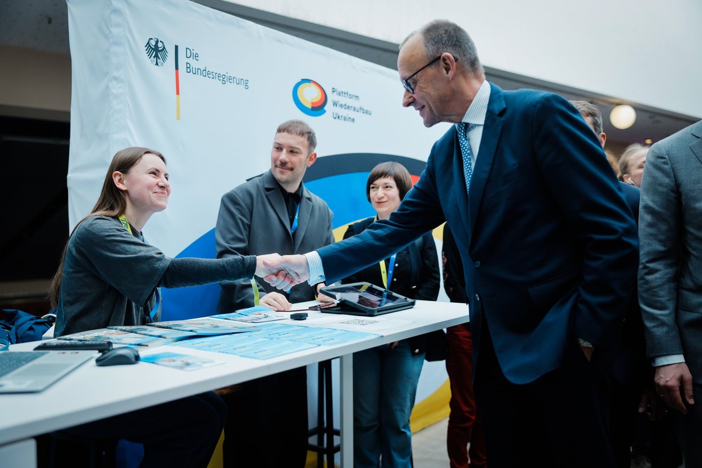 Foto von einem stehenden Mann, der einer sitzenden Frau die Hand schüttelt.