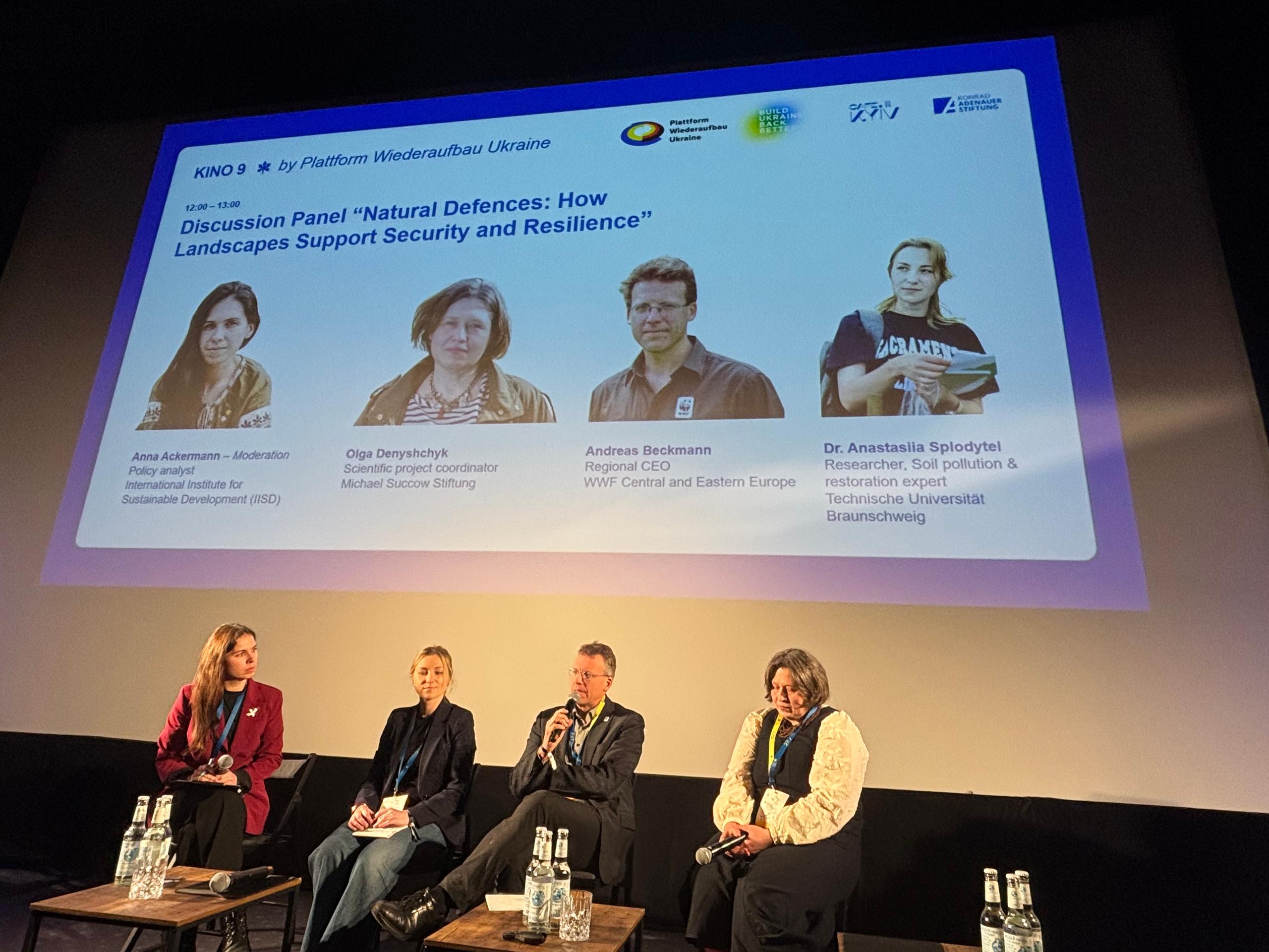 Foto von vier Menschen auf einem Panel, im Hintergrund einer Präsentation.