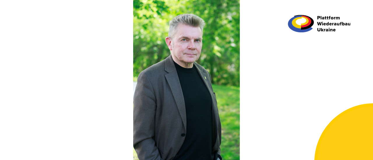 Mann mit grauem Sakko und schwarzem Shirt vor grünem Hintergrund.